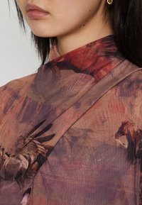 Top trasparente con una stampa astratta di cavalli in tonalità calde di rosso, rosa e marrone, dotato di un colletto alto e tessuto drappeggiato.