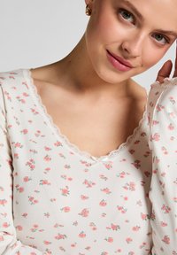 Long-sleeve top in soft white ribbed fabric with a floral print in pink and green; features a lace-trimmed V-neck and delicate accents.