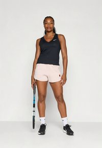 Top atlético preto e calções rosa claro, em pé com uma raquete de ténis. Apresenta um design em decote em V e um logótipo no top.