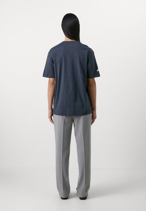 Persoon met lang, donker haar staat met de rug naar de camera, draagt een donkerblauw shirt met korte mouwen en lichtgrijze broek tegen een effen achtergrond.