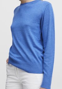 Femme portant un pull bleu ajusté à manches longues associé à un pantalon blanc, main posée de manière décontractée dans la poche, sur un fond blanc uni.