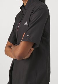 Veste noire à manches courtes avec col montant, dotée d'une fermeture éclair à l'avant, d'un accent rose et d'un logo réfléchissant sur la poitrine. Texture lisse.