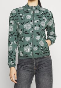 Grön zip-jacka med blommönster, med svarta blad och vita blommor. Ribbad krage och manschetter, texturerad tyg. Bärs med mörka jeans.