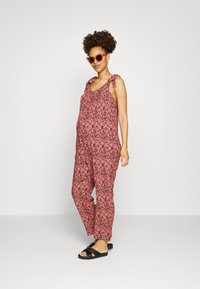 Macacão rosa com padrão, de corte largo, alças ajustáveis e sandálias pretas. Apresenta um design floral e tecido suave. Modelo posando contra um fundo branco.