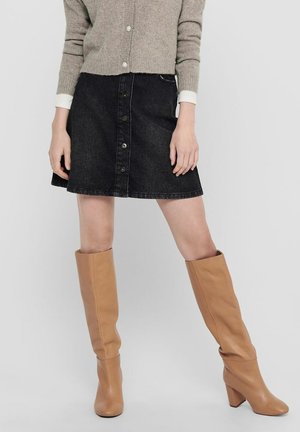 Kobieta ubrana w beżowy sweter na guziki, czarną dżinsową spódnicę na guziki z przodu oraz brązowe wysokie botki na obcasie, stojąca z lekko rozstawionymi nogami.