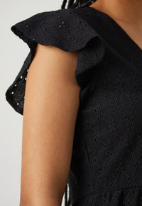 Vestido preto com mangas franzidas e tecido texturizado com padrões de ilhós, destacando um design intrincado na área dos ombros.