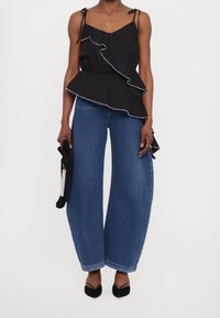 Frau trägt ein schwarzes, rüschengesäumtes ärmelloses Oberteil, weit geschnittene blaue Jeans, schwarze spitz zulaufende Pumps und hält eine schwarze strukturierte Clutch-Tasche.