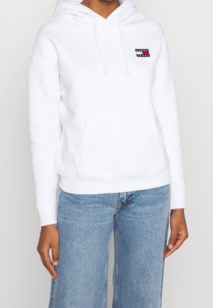 Person trägt einen weißen Hoodie mit Fronttasche und Tommy Jeans Logo auf der Brust, kombiniert mit hellblauen Jeans aus Denim.
