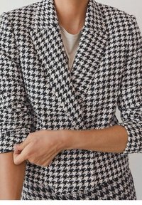 Personne portant un blazer pied-de-poule noir et blanc en train de retrousser une manche, avec un haut beige clair en dessous.