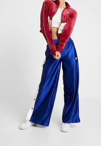 Röd sammetsjacka med dragkedja och vita detaljer, tillsammans med blå satinställbyxor med vita tryckknappar och guldlogga. Vita sneakers fulländar looken.