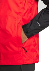 Main dans la poche d'une veste rouge avec manche noire étiquetée « DRYVENT », montrant une sangle de poignet réglable et un cordon de serrage à l'ourlet.