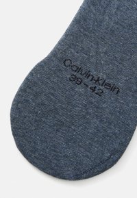 Siva-modra nogavica iz teksturiranega blaga. Na območju prstov je v črni barvi izvezeno ime "Calvin Klein" in velikost "38-42".