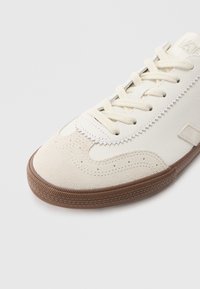 Bijela kožna tenisica s taupe antilop akcentima, okruglim vrhom, ravnim vezicama i teksturiranim smeđim gumiranim đonom s uzorkom gaznoga dijela.