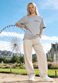 Jeune femme en pantalon beige et t-shirt gris se tient dans un jardin avec une grande roue et des bâtiments classiques en arrière-plan sous un ciel bleu.