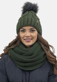Dark green knitted beanie with a fur pom-pom and matching thick scarf featuring a textured pattern, both made from warm, cozy material.