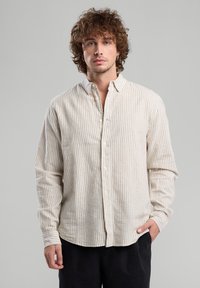 Camicia a righe crema e beige, design con bottoni, maniche lunghe, texture morbida, orlo curvo, piccolo dettaglio del logo vicino alla pattina dei bottoni.