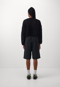 Pull court en maille noire avec une structure ample, associé à un short noir et des chaussures de sport avec accents verts. Chaussettes blanches visibles.