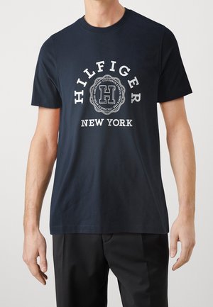 Mężczyzna noszący czarną koszulkę z krótkim rękawem z białym napisem "HILFIGER NEW YORK" i okrągłą emblematem na środku klatki piersiowej.
