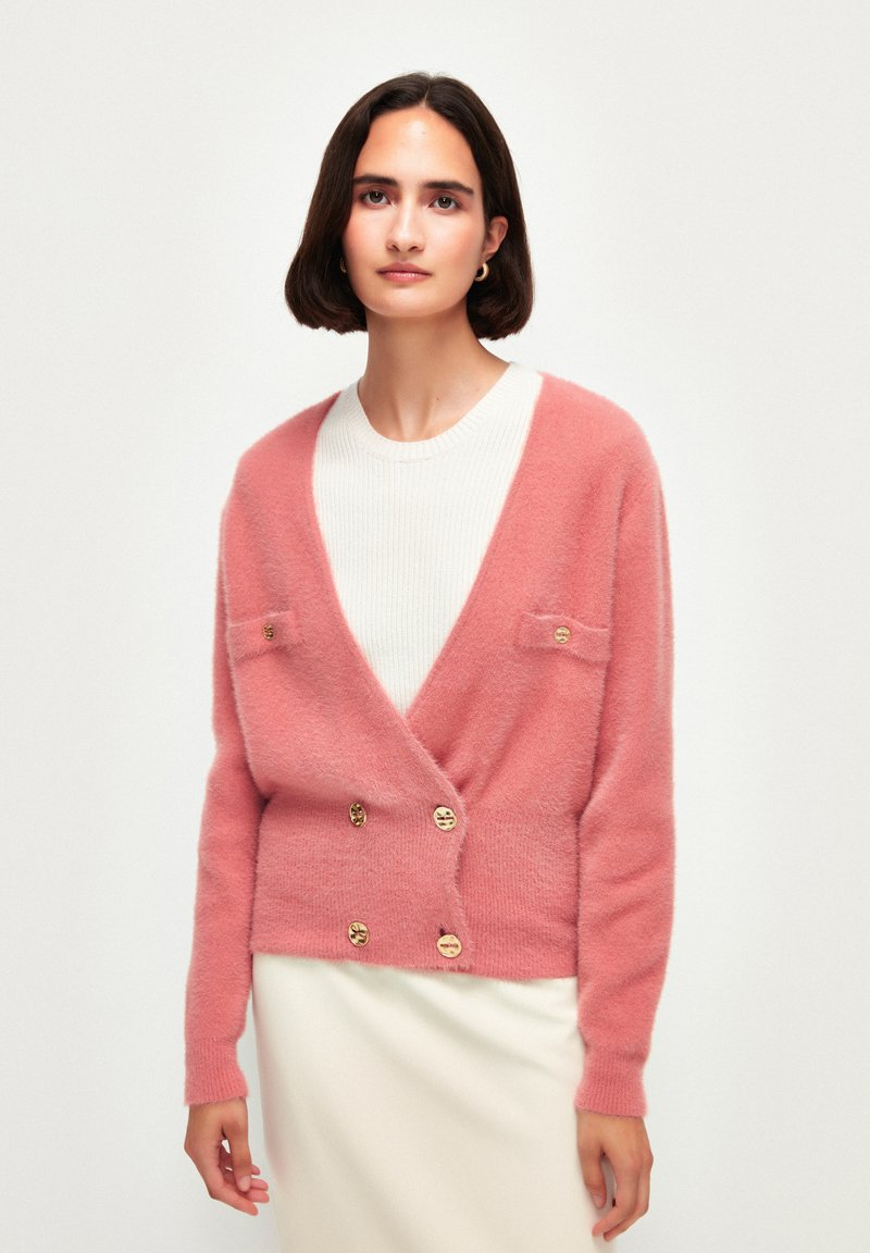 Cardigan rosa fluffy con scollatura a V, design doppiopetto, due tasche frontali e bottoni dorati, indossato sopra una maglia bianca texturizzata.