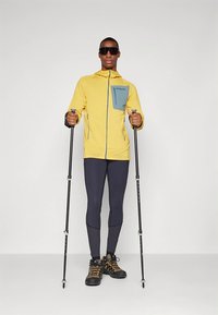 Gul zip-up jacka med blå bröstficka, svarta leggings och svarta vandringsskor. Modellen håller vandringsstavar och bär solglasögon.