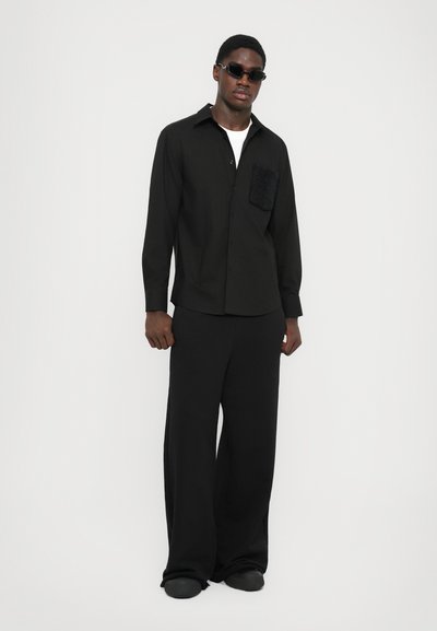Homme portant des lunettes de soleil noires, une chemise noire à boutons avec une poche texturée sur la poitrine, un pantalon noir ample et des chaussures noires, se tenant contre un fond blanc.