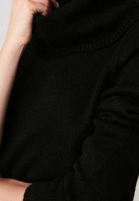 Person, der den Kragen eines schwarzen, strukturierten Langarmpullovers anpasst, gezeigt von den Schultern bis zur Mitte des Oberkörpers.