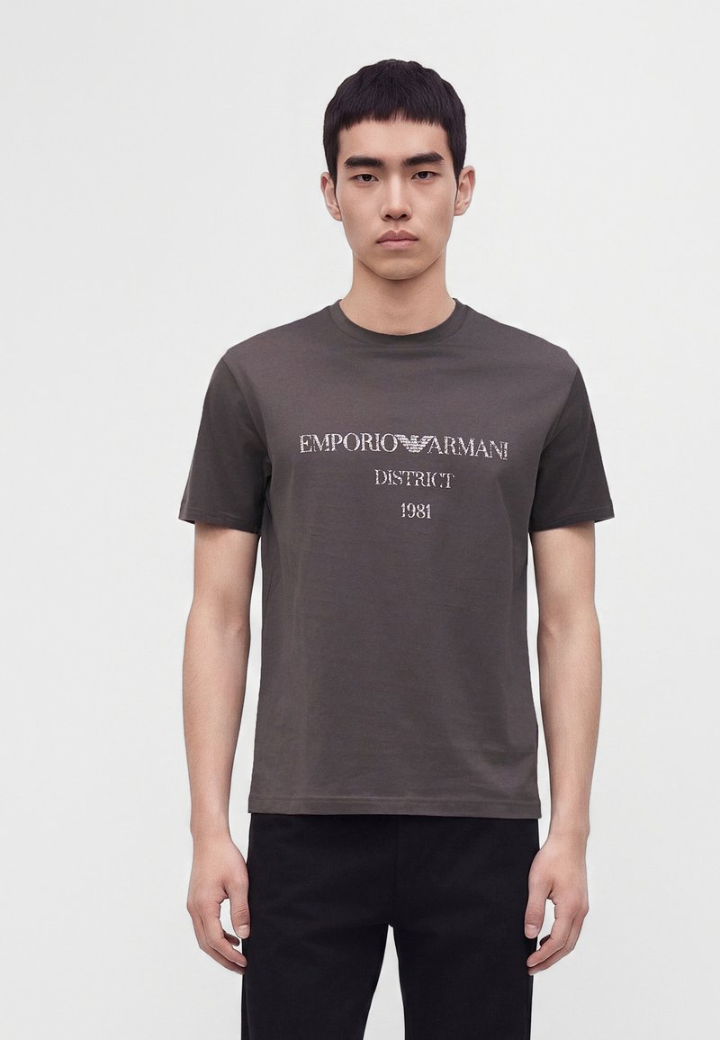 Νέος άνδρας που φοράει ένα σκούρο γκρι T-shirt Emporio Armani με το κείμενο "District 1981", στέκεται μπροστά σε ένα απλό φωτεινό φόντο.