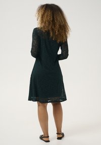Robe en dentelle vert foncé à manches longues, avec un motif floral texturé, un ourlet au niveau du genou et une coupe décontractée. Sandales plates noires visibles.