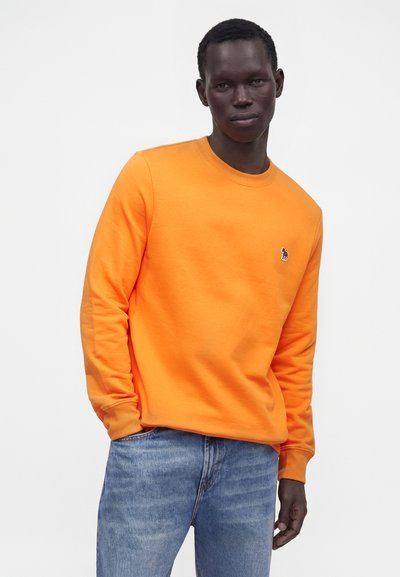 Jeune homme portant une chemise orange à manches longues et un jean bleu, debout devant un fond clair uni.