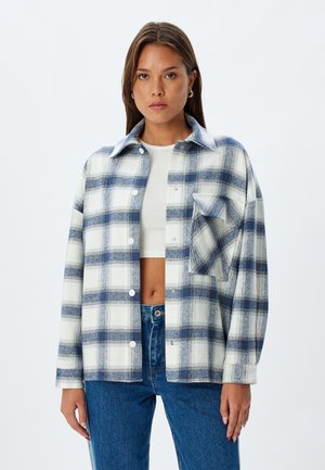 Plaid button-up shirt in blue and white, made of fabric with a soft texture. Features a collar and a chest pocket. Worn over a white top.