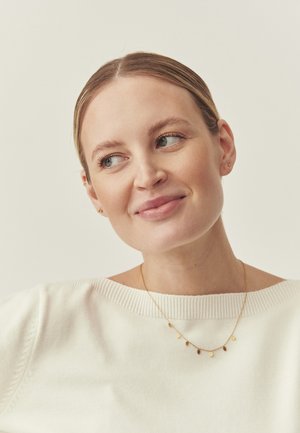 Auksinė kaklo grandinėlė su keliomis mažomis apvalios ir ovalios formos pakabukais, dėvima ant baltos briaunuotos megztinio, pasižyminčio minkšta audinio tekstūra.