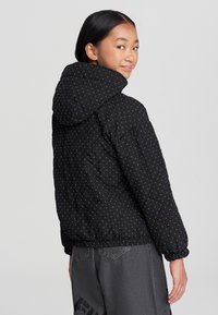 Giubbotto nero trapuntato con cappuccio, caratterizzato da un motivo a pois, polsini elastici e orlo arricciato. Texture liscia con una silhouette moderna.