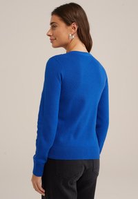 Zila adīta džemperis ar teksturētu rakstu, apaļu dekoltē un ribotām piedurknēm. Nēsāts kopā ar melnām biksēm, rādot pielāgotu siluetu no aizmugures.