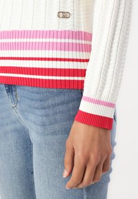 Maglione a costine bianco con righe orizzontali rosa e rosse, caratterizzato da un dettaglio con gancio argentato sul petto e polsini a contrasto.