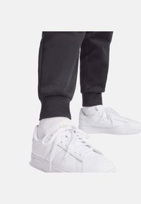 Scarpe da ginnastica bianche con lacci e tre strisce laterali, abbinate a pantaloni da tuta neri con risvolto e calzini bianchi alla caviglia.