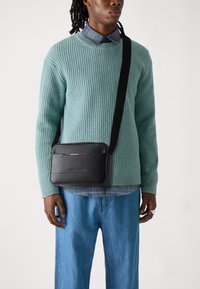 Sac en cuir noir texturé avec une forme plate et une bandoulière réglable, porté en travers du corps sur un pull côtelé bleu clair et une chemise à carreaux.