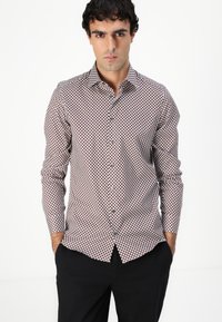 Chemise blanche à boutons avec un motif géométrique rouge et marine, à manches longues et avec un col standard, associée à un pantalon sombre.