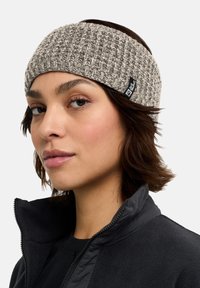 Bandeau tricoté en gris clair avec un motif texturé. Présente une étiquette noire. Porté avec une veste en polaire noire. Tissu doux, ajustement serré.