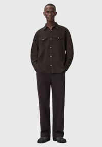 Μαύρο πουκάμισο με κουμπιά και δύο τσέπες στο στήθος, συνδυασμένο με μαύρο παντελόνι. Το πουκάμισο διαθέτει γιακά και είναι κατασκευασμένο από αδιαφανές ύφασμα.