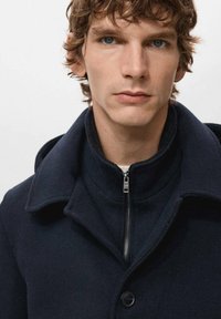 Abrigo de lana azul marino con cierre de cremallera y cuello ancho. Presenta un botón y una textura sutil, ofreciendo una silueta moderna y ajustada.