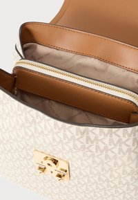 Płócienna torba w kolorze kremowym z geometrycznym wzorem logo. Posiada zapięcie ze złotym okuciem, brązowe skórzane akcenty oraz zapinaną na suwak wewnętrzną kieszeń.