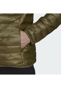 Main dans la poche d'une veste matelassée vert olive avec des coutures horizontales et un poignet de manche élastiqué.