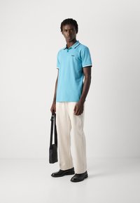 Άνδρας φορώντας ένα ανοιχ�τό μπλε πόλο μπλουζάκι, κρέμ παντελόνι, μαύρα παπούτσια, κρατώντας μια μικρή μαύρη τσάντα ώμου μπροστά σε λευκό φόντο.