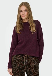 Burgunderfarbener Strickpullover mit Rundhalsausschnitt; kombiniert mit braunen Jogginghosen mit Leopardenmuster, die eine lockere Passform und eine strukturierte Oberfläche haben.