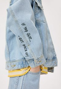 Giacca in denim azzurro chiaro con vestibilità rilassata, chiusura a bottoni e testo ricamato nero sulla manica sinistra. Design minimalista.