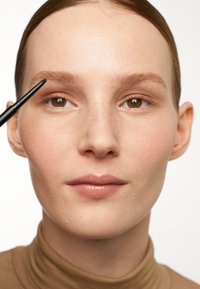 Model met een lichte huid, gekleed in een bruine coltrui. Oogpotlood in de hand, gefocust op gedefinieerde wenkbrauwen. Gladde textuur, natuurlijke make-up.