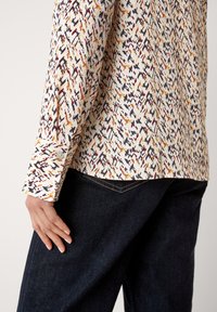 Camicia a maniche lunghe con un motivo astratto multicolore su sfondo crema, abbinata a jeans di denim scuro. È visibile il dettaglio del polsino con bottone.