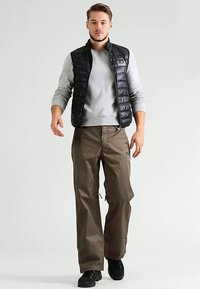 Czarna pikowana kamizelka na jasnoszarej bluzie, zestawiona z luźnymi brązowymi spodniami i czarnymi butami; cechy charakterystyczne to boczne kieszenie i zamki błyskawiczne.