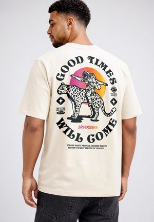 Mann trägt ein beiges T-Shirt mit einer Grafik eines Astronauten, der auf einem gefleckten Leoparden reitet, und dem Text „Good times will come“ auf dem Rücken.