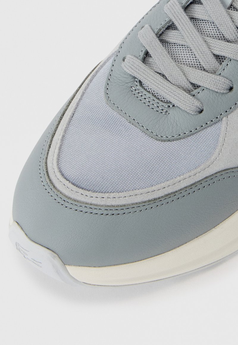 Gros plan sur l'orteil d'une chaussure de sport gris clair avec des couches de tissu, des panneaux en daim et en cuir, et des lacets gris assortis sur une semelle blanche.